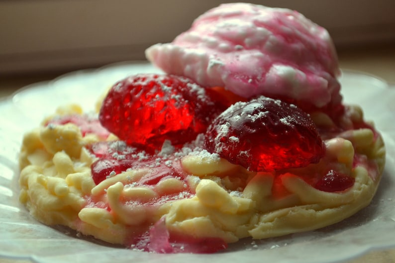 May include: A white cookie shaped like a pretzel with red and pink jelly candies on top. The cookie is covered in powdered sugar.