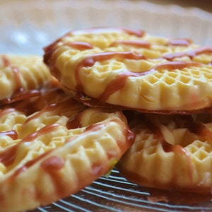 Könnte beinhalten: Nahaufnahme von mehreren runden Keksen mit Waffelstruktur, beträufelt mit einer rotbraunen Glasur. Die Kekse sind auf einem klaren Glasteller gestapelt und bilden eine ansprechende Anordnung. Die Kekse haben eine goldgelbe Farbe.