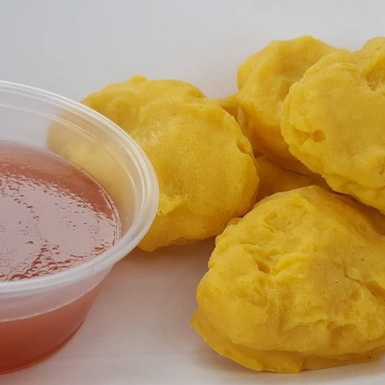 K&ouml;nnte beinhalten: Eine Gruppe goldgelber, abgerundeter, frittierter Lebensmittel, die neben einem kleinen, durchsichtigen Plastikbeh&auml;lter mit einer glatten, r&ouml;tlich-orangen Dip-Sauce angeordnet sind. Die Speisen haben eine texturierte Oberfl&auml;che und eine gelbe Farbe.
