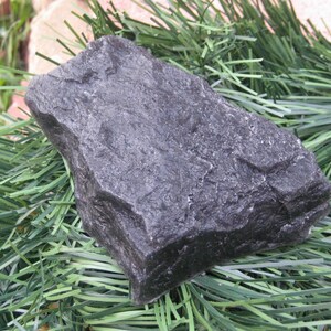 May include: A dark gray, irregularly shaped rock with a rough, textured surface. The rock is sitting on a bed of green artificial pine needles.