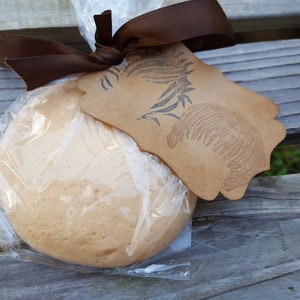 May include: A round, light brown baked good wrapped in clear cellophane, tied with a brown ribbon, and a decorative tag. The tag has a leaf and shell design. The item is on a wooden surface.