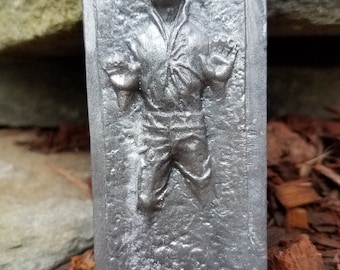 May include: A silver-colored, rectangular, 3D printed replica of Han Solo frozen in carbonite. The replica is detailed with a textured surface and a raised image of Han Solo.