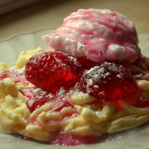May include: A white and pink meringue dessert topped with red jelly candies and powdered sugar. The dessert is on a clear plate.