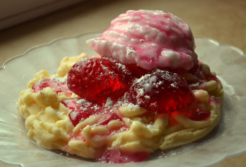 May include: A white and pink meringue dessert topped with red jelly candies and powdered sugar. The dessert is on a clear plate.