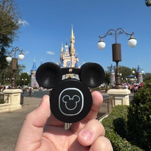 May include: A black Mickey Mouse-shaped accessory with a white outline of Mickey's face on a circular base. The accessory has two round ears and is held in front of a castle on a sunny day.