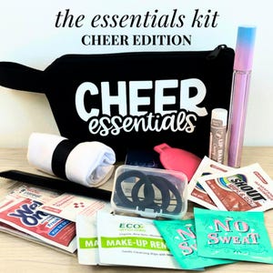 May include: A black zippered pouch with the white text "CHEER essentials" containing a white towel, a black comb, a clear case with black hair ties, a pink hair clip, a lip balm, a tall ombre tube, and various single-use packets labeled "SHOUT", "NO SWEAT", and "MAKE-UP REMOVER".