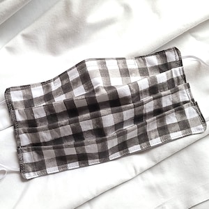 May include: A handmade face mask with a black and white gingham pattern. The mask is made of fabric and has elastic ear loops. The mask is folded and has multiple pleats. The background is a white fabric.