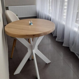 May include: A circular wooden table with a light-coloured top and a white, starburst-shaped base. A beige chair is positioned next to the table. Sheer white curtains frame a window in the background.