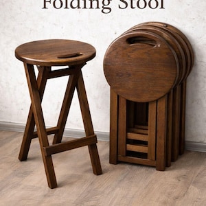Könnte beinhalten: Dunkler Holz-Klapphocker mit rundem Sitz und Griffaussparung. Der Hocker ist dunkelbraun und hat ein faltbares Design. Mehrere zusammengeklappte Hocker sind aufeinander gestapelt. Der Text "Dark Wood Folding Stool" steht oben.