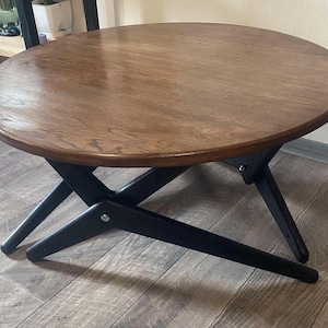 Puede incluir: Una mesa de centro redonda con una tapa de madera oscura y patas negras en ángulo. La mesa tiene un perfil bajo y un diseño moderno de mediados de siglo. El diámetro de la mesa es de aproximadamente 91 cm.