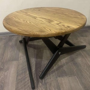 May include: A round wooden coffee table with a light brown, wood-grain top and black legs. The legs have a unique, angled design. The table is approximately 76 cm in diameter and 41 cm high.