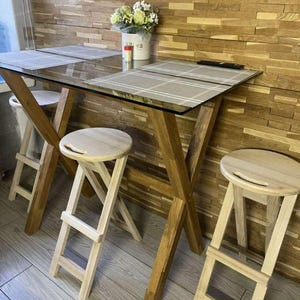 May include: A contemporary dining set comprising a rectangular glass-topped table with wooden X-frame legs. Two matching wooden stools are placed in front of the table. A vase of flowers sits on the table, with placemats.