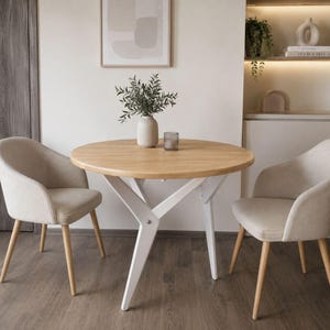 Puede incluir: Una mesa de comedor redonda de madera con patas blancas y dos sillas tapizadas beige. La mesa está coronada con un jarrón de vegetación y un pequeño portavelas de cristal. La mesa tiene unos 100 cm de diámetro.