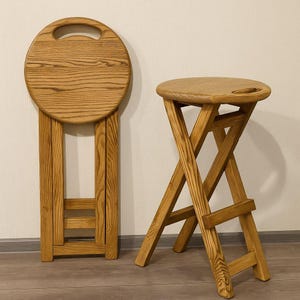 Op de afbeelding: Twee houten klapstoelen. Eén is opgevouwen, met een ronde zitting met een handvat en rechthoekige poten. De andere is open, met een X-vormig pootontwerp en een ronde zitting met een handvat. Beide zijn gemaakt van lichtgekleurd hout.
