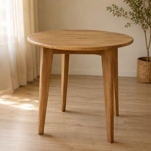以下が含まれることがあります： 明るい茶色の仕上げの丸い木製テーブル。テーブルには4つの角度の付いた脚と滑らかな天板があります。テーブルは自然光のある部屋にあり、背景には植物があります。テーブルの直径は約76cmです。