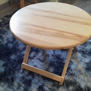 May include: A round wooden table with a natural wood finish and a foldable design. The table is sitting on a blue and white fluffy rug.