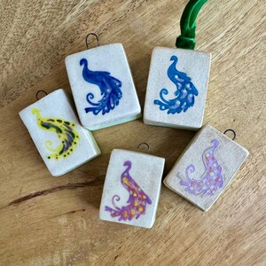 Peut inclure: Cinq ornements rectangulaires en c&eacute;ramique avec des motifs de paon de diff&eacute;rentes couleurs. Chaque ornement est dot&eacute; d'une boucle m&eacute;tallique pour l'accrochage. Les couleurs du paon comprennent le bleu, le jaune, le violet et un ruban vert sur un ornement.