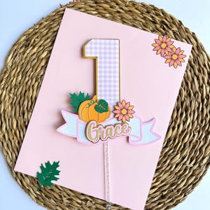 May include: A pink card with a gingham number one cake topper. The topper has a pumpkin, flowers, and a banner that says "Grace". The card also has green leaves and flowers.
