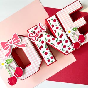 May include: A layered papercraft design spelling "ONE" with a bow, cherries, and the word "Sweet". The letters are red and white, with a gingham pattern on the "O" and a cherry pattern on the "N" and "E". A daisy is next to the "E".