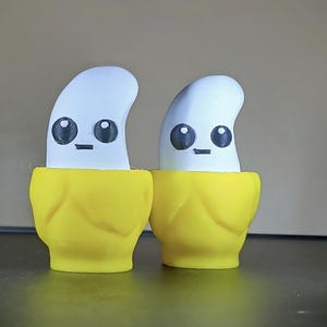 May include: Two whimsical banana-shaped figurines. Each has a white, curved top with black eyes and a small mouth, set in a bright yellow peel. The figures are side-by-side, creating a playful and cheerful aesthetic.