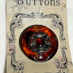 May include: A large, round button with a dark amber and brown marbled appearance. The button has a raised, cross-shaped center and is mounted on a vintage-style card that reads "BUTTONS" in elegant script, framed by floral designs.
