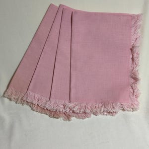 May include: Four pink linen napkins with fringed edges are fanned out on a white surface. The napkins are a solid, soft pink colour. The fringed edges add a decorative touch to the napkins.