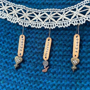 Pode incluir: Brincos feitos à mão com etiquetas de madeira e amuletos decorativos. As etiquetas têm as palavras "shine", "inspire" e "shine" escritas nelas. Os brincos penduram-se de uma guarnição de renda branca contra um fundo azul.