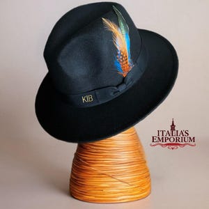Puede incluir: Un sombrero fedora negro con un acento de pluma colorida. El sombrero tiene una cinta negra con las iniciales "KIB" en dorado. El sombrero se exhibe en un soporte de madera. La pluma es naranja, azul y verde.