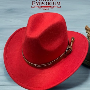 Puede incluir: Un sombrero de vaquero rojo brillante con una banda y hebilla de cuero marrón. El sombrero tiene un ala ancha y una forma clásica. El texto "ITALIA'S EMPORIUM" es visible en la parte superior de la imagen.