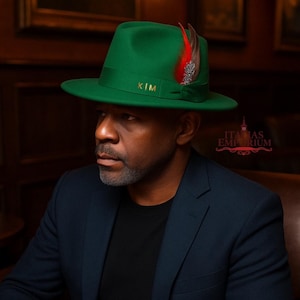 Puede incluir: Un sombrero fedora verde con una pluma decorativa y las iniciales "KIM" en dorado. El sombrero tiene un ala ancha y una cinta de grogrén. El sombrero se lleva con una blazer azul marino.