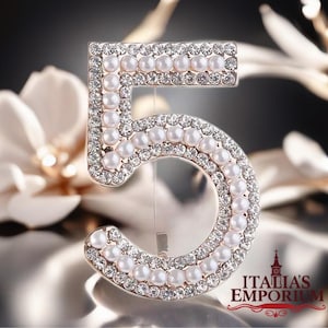 May include: A decorative number 5 brooch, embellished with faux pearls and clear crystals. The brooch has a silver-tone metal frame and a pin on the back. The background is blurred, with a soft, neutral color palette.