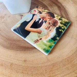 May include: A square ceramic coaster with a printed image of a couple embracing in a green outdoor setting. The couple is dressed in formal attire, with the woman wearing a white dress and the man wearing a black suit.