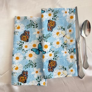 Puede incluir: Dos servilletas de tela azules dobladas con un estampado floral y de mariposas. Las servilletas presentan margaritas blancas, hojas verdes y mariposas monarca naranjas y negras. Un cuchillo y una cuchara se colocan junto a las servilletas.