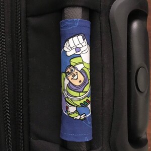 May include: A blue luggage handle cover with a cartoon image of Buzz Lightyear from the Toy Story movie franchise. The cover is made of fabric and has a white zipper.