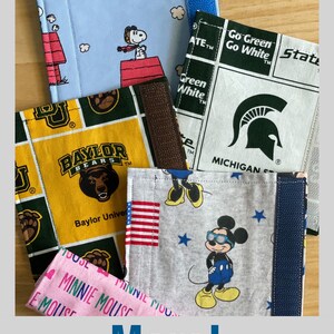 May include: Five different fabric swatches with various designs. The swatches are folded and stacked on top of each other. The top swatch is light blue with a white cloud pattern and a cartoon Snoopy. The second swatch is white with a green and white checkered pattern and the text "Go Green Go White". The third swatch is yellow with a green and yellow checkered pattern and the text "Baylor Bears". The fourth swatch is gray with a white star pattern and a cartoon Mickey Mouse wearing sunglasses. The fifth swatch is pink with a white heart pattern and the text "Minnie Mouse".