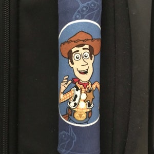 May include: Blue luggage handle cover with a cartoon image of Woody from Toy Story. The cover has a white outline around Woody and a blue background with a silhouette of the spaceship from Toy Story.