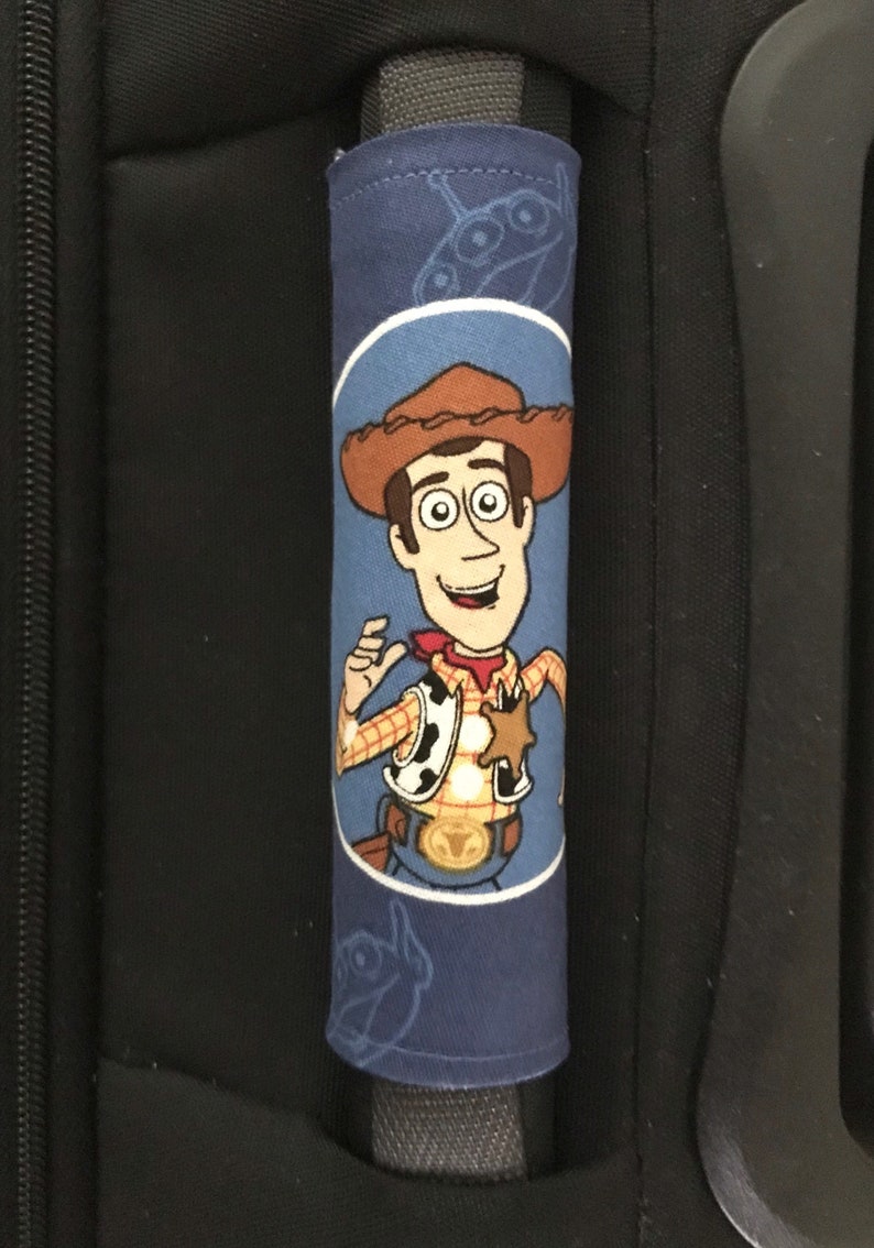 May include: Blue luggage handle cover with a cartoon image of Woody from Toy Story. The cover has a white outline around Woody and a blue background with a silhouette of the spaceship from Toy Story.