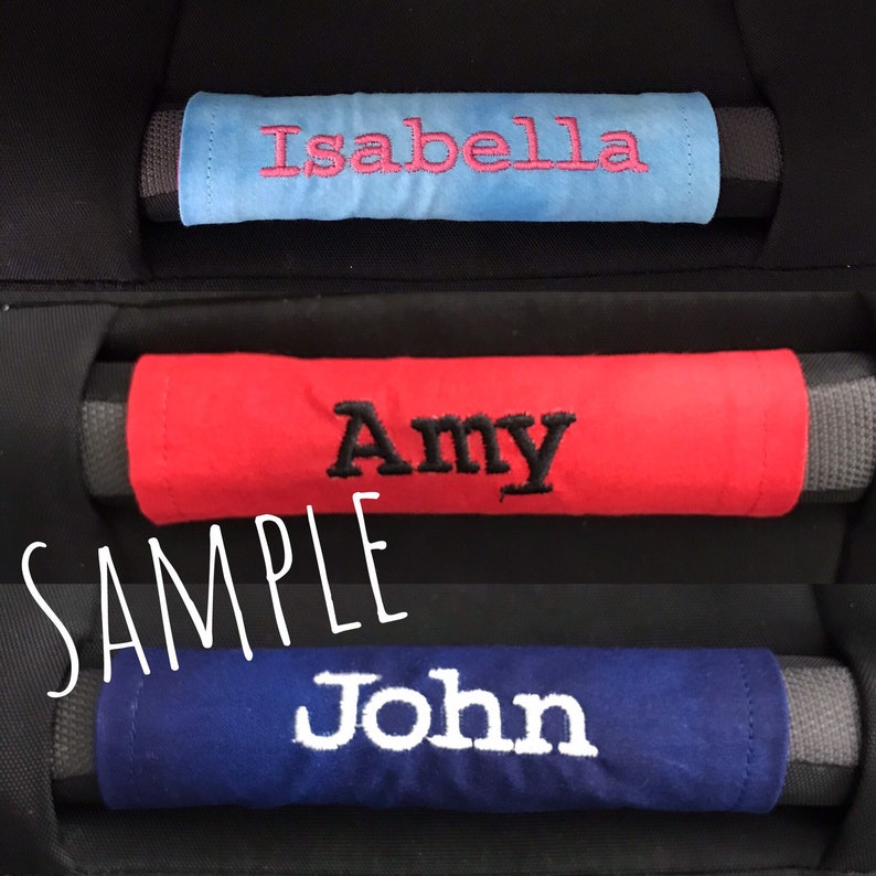 May include: Three fabric car seat belt covers with embroidered names. The top cover is light blue with the name "Isabella" embroidered in pink. The middle cover is red with the name "Amy" embroidered in black. The bottom cover is dark blue with the name "John" embroidered in white.