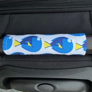 May include: A black suitcase with a blue and yellow patterned handle cover featuring a cartoon fish design. The handle cover is made of fabric and has a white background.