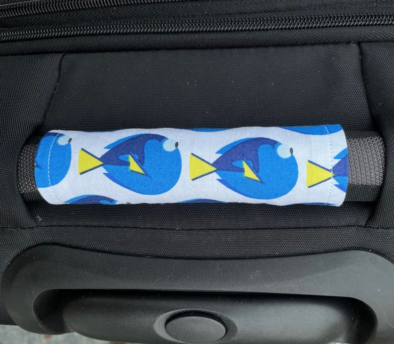 May include: A black suitcase with a blue and yellow patterned handle cover featuring a cartoon fish design. The handle cover is made of fabric and has a white background.