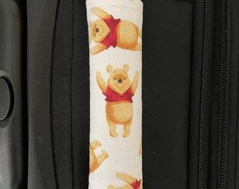 May include: A white luggage handle cover with a repeating pattern of Winnie the Pooh. The cover is made of fabric and has a sewn seam around the edges.