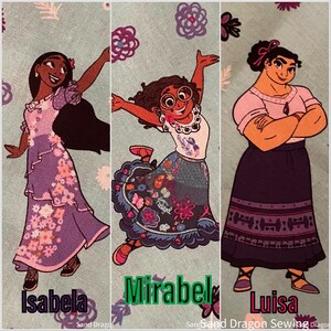 May include: Three cartoon characters from the movie Encanto.  The characters are Isabela, Mirabel, and Luisa.  Isabela is wearing a purple dress with floral designs.  Mirabel is wearing a blue and white dress with floral designs.  Luisa is wearing a purple skirt with a red and white pattern and a pink shirt.