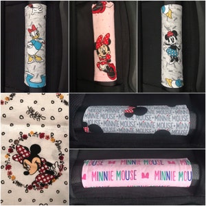 May include: Set of five car seat belt covers featuring different Minnie Mouse designs. The covers are made of fabric and have a variety of colors and patterns, including pink, white, black, and red. One cover features a Minnie Mouse with a red bow and polka dot dress, another features a Minnie Mouse with a pink bow and polka dot dress, and another features a Minnie Mouse with a pink bow and a white dress. The covers are all approximately 10 inches long and 3 inches wide.