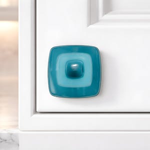 May include: A teal square cabinet knob with a central, raised square detail. The knob is mounted on a white cabinet door. The design is modern and minimalist, suitable for kitchen or bathroom cabinetry.