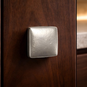 May include: A square, brushed nickel cabinet knob is mounted on a dark brown wooden cabinet door. The knob has a textured surface and reflects light. The cabinet door has a rich, wood grain appearance.