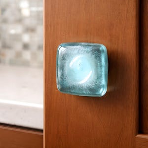 May include: A square, light blue glass cabinet knob with a frosted interior, mounted on a wooden cabinet door. The knob has a slightly rounded shape and is set against a warm, brown wood grain.