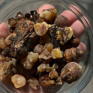 May include: A close-up of a small bowl filled with various sizes and shapes of amber-colored resin. The resin is a mix of light and dark brown colors, with some pieces having a translucent quality.