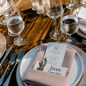 May include: A place setting with a white plate, a pink napkin, and a menu card with the text "Clare & Jared" and "Main Course". The menu card also includes the text "Steve Doerffler".