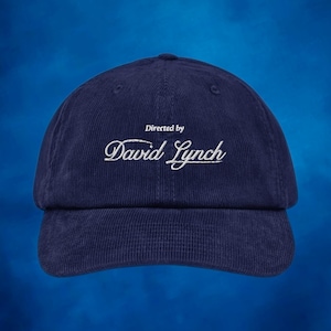 Peut inclure: Une casquette de baseball en velours côtelé bleu marine avec une broderie blanche qui dit "Directed by David Lynch".