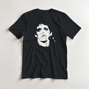 Puede incluir: Camiseta negra con un gráfico blanco de un rostro. El gráfico es un retrato estilizado con cabello oscuro y una expresión concentrada. La camiseta es de estilo cuello redondo clásico.